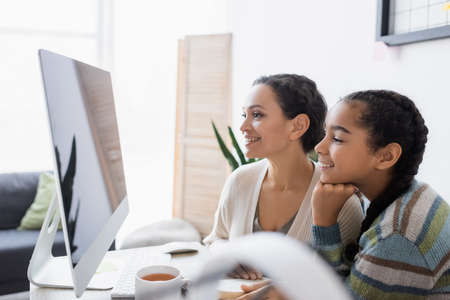 cheerful african american mother and daughter looking at computer monitor while doing homework togetherの写真素材
