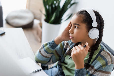 thoughtful african american teen girl in headphones holding pen and looking away while studying at...