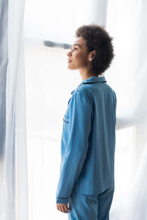 Side view of african american woman in blue pajamas standing near curtainsの写真素材