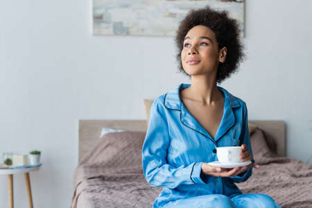 Young african american woman in pajamas holding white cup and saucer in bedroom