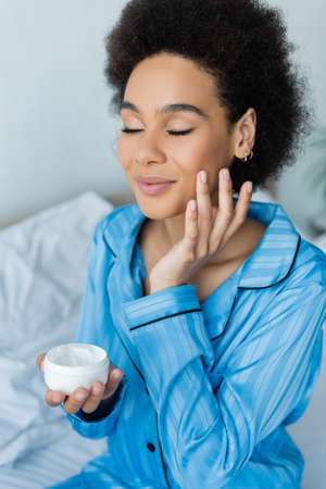 smiling african american woman in pajamas holding container and applying face cream in bedroomの写真素材