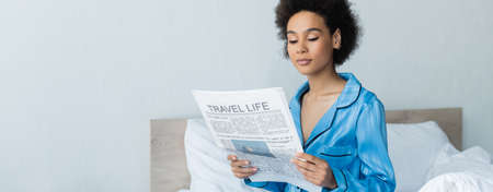 african american woman in pajamas reading travel life newspaper in bedroom, bannerの写真素材