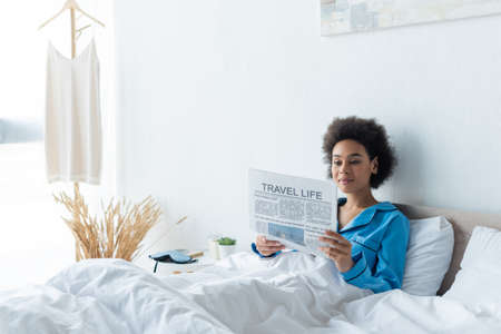 young african american woman in pajamas reading travel life newspaper in bedの写真素材