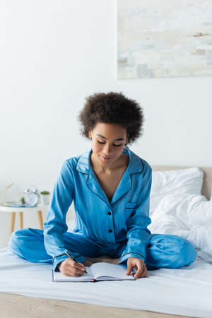 curly african american woman in pajamas sitting on bed and writing on notebookの写真素材