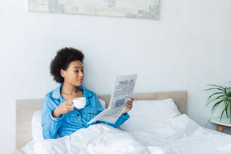 african american woman in pajamas reading travel life newspaper while holding cup of coffee in bedroomの写真素材