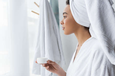 side view of african american woman in bathrobe holding container with face cream in bathroomの写真素材