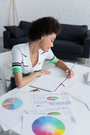 Side view of african american woman writing on notebook near documents with graphs at homeの写真素材