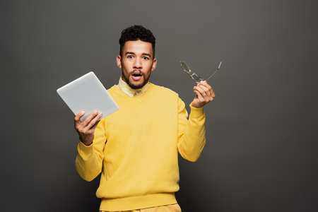 shocked african american man holding glasses and digital tablet on dark grey