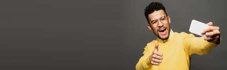 young excited african american man in glasses taking selfie isolated on grey, banner