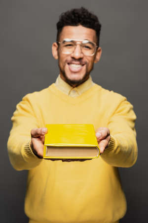 cheerful and blurred african american man in glasses and yellow outfit holding book on dark greyの写真素材