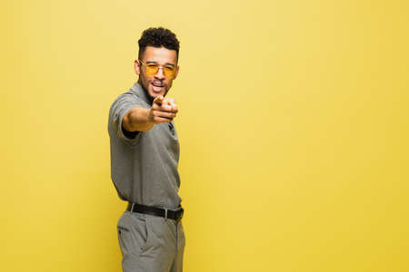 positive african american man in sunglasses and grey tennis shirt pointing at camera isolated on...