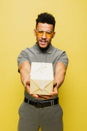 blurred african american man in sunglasses and grey tennis shirt holding wrapped present isolated on yellowの写真素材