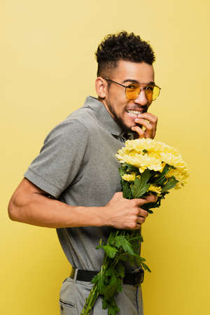 worried african american man in sunglasses and grey tennis shirt holding bouquet of flowers isolated on yellowの写真素材