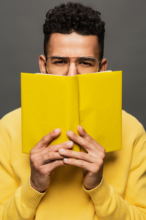 african american man in glasses and yellow outfit holding book isolated on greyの写真素材