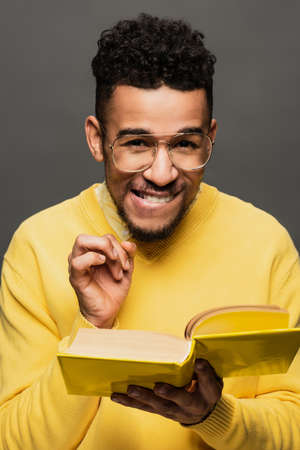 sly african american man in glasses and yellow sweater holding book and smiling isolated on grey