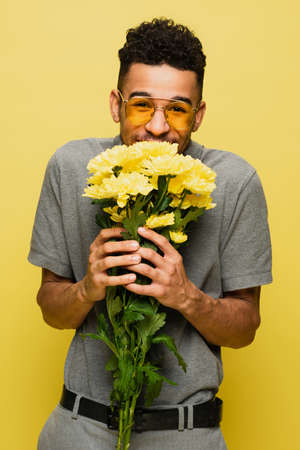 happy african american man in sunglasses and grey tennis shirt holding bouquet of flowers isolated on yellowの写真素材
