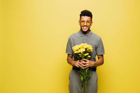 happy african american man in sunglasses and grey tennis shirt holding bouquet of flowers isolated on yellowの写真素材
