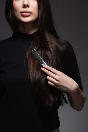 partial view of young woman brushing long shiny hair with comb isolated on dark grey