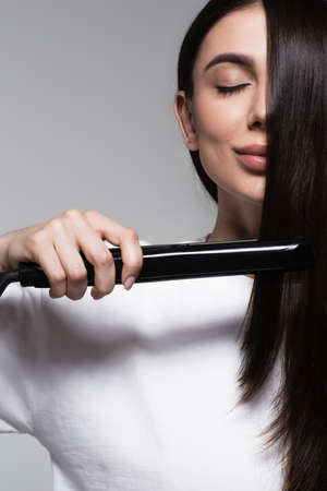 young and pleased woman using hair straightener isolated on greyの写真素材