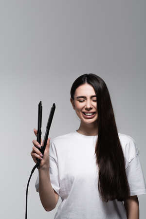 happy brunette woman holding hair straightener isolated on greyの写真素材