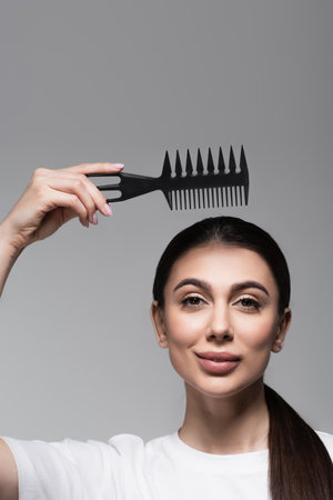 cheerful woman in white t-shirt holding comb above straight hair isolated on greyの写真素材