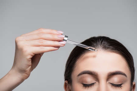 cropped view of young woman holding pipette with oil near shiny hair isolated on greyの写真素材