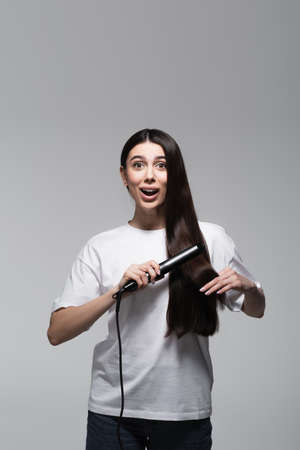 young amazed woman using hair straightener isolated on greyの写真素材