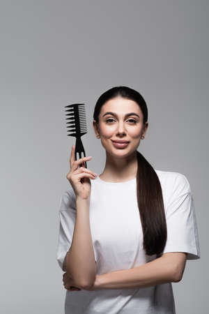 happy woman in white t-shirt holding comb near straight hair isolated on greyの写真素材
