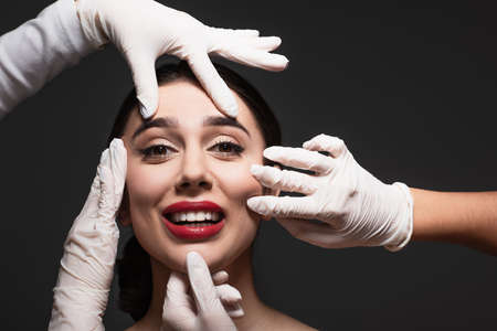 plastic surgeons in latex gloves touching face of smiling young woman isolated on blackの写真素材