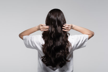 back view of brunette woman adjusting long shiny hair isolated on greyの写真素材