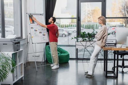 full length of african american man pointing at charts on flip chart near blonde colleague