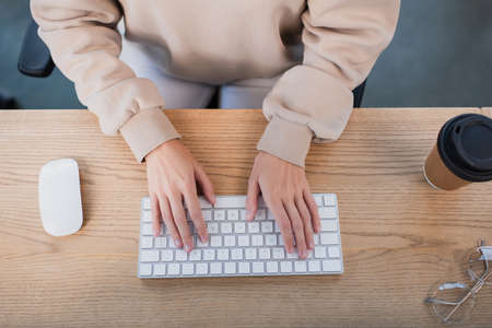top view of businesswoman typing on computer keyboard near paper cup and glassesの写真素材