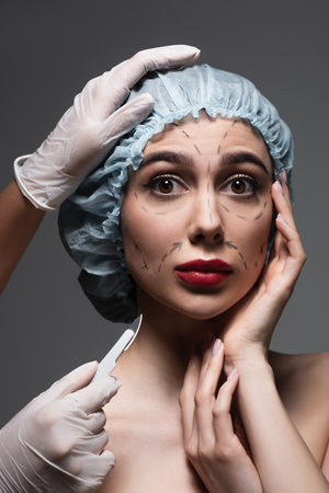 plastic surgeon in latex gloves holding scalpel near scared woman with marked lines on face isolated on dark greyの写真素材