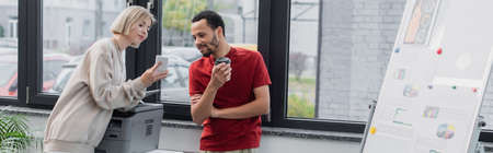 blonde woman showing smartphone to african american colleague with paper cup near printer, banner