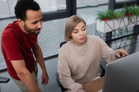 high angle view of blonde manager pointing at computer monitor near african american colleague