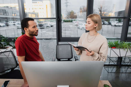blonde businesswoman holding notebook near computer monitor and african american colleagueの写真素材