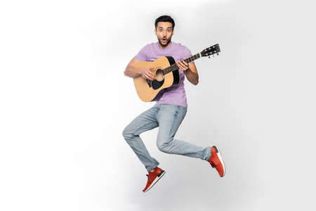 shocked man in jeans and purple t-shirt levitating while playing acoustic guitar on white