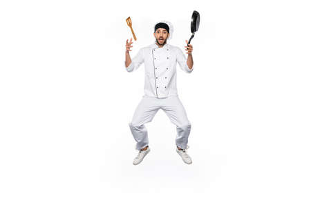 shocked chef in hat and uniform levitating near flying frying pan with wooden spatula isolated on...