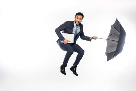 astonished businessman in suit jumping with laptop and umbrella on white