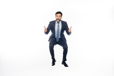excited businessman in suit jumping while showing thumbs up on white