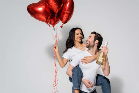 man holding champagne bottle while piggybacking cheerful woman with heart-shaped balloons on greyの写真素材