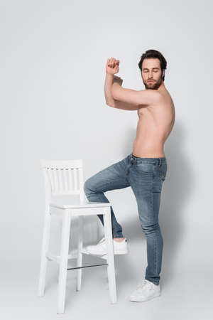 full length view of shirtless man in jeans posing near white chair on greyの写真素材