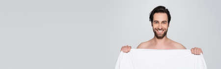 cheerful shirtless man looking at camera while holding white blanket isolated on grey, bannerの写真素材