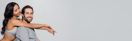 young man smiling at camera near seductive woman in bra embracing him isolated on grey, bannerの写真素材