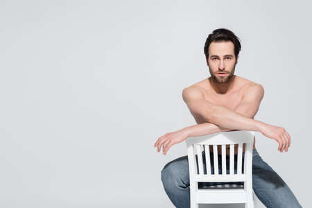 young and sexy shirtless man sitting on white chair and looking at camera on greyの写真素材