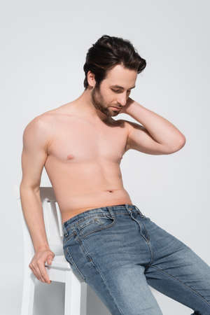 shirtless man in jeans touching neck while sitting on white chair on greyの写真素材