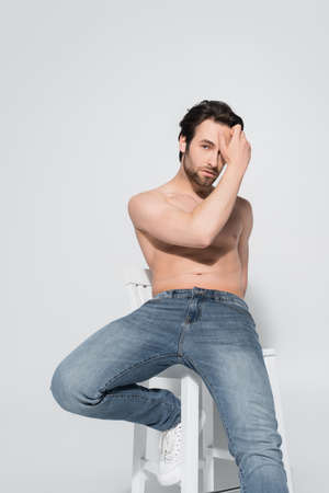 brunette shirtless man in jeans looking at camera while holding hand near face on greyの写真素材
