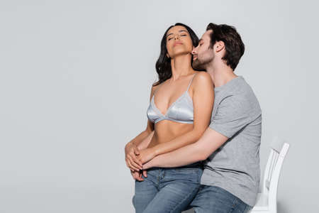 man in t-shirt hugging passionate woman in satin bra while sitting on chair on greyの写真素材