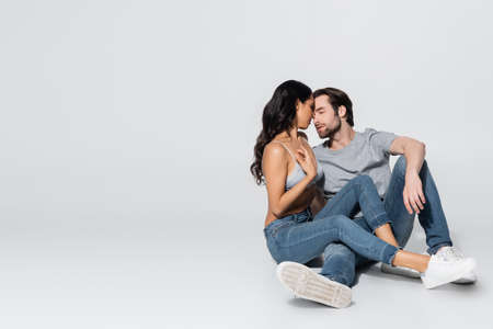 sexy brunette woman in jeans and satin bra seducing young man while sitting on grey