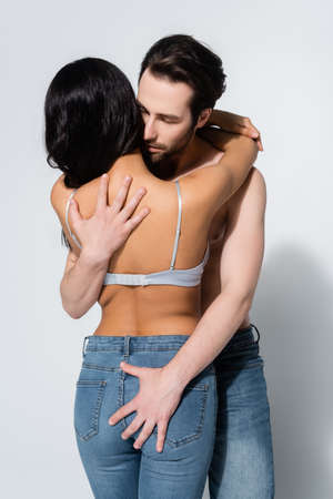 back view of passionate woman in jeans and bra near young shirtless man hugging her on greyの写真素材